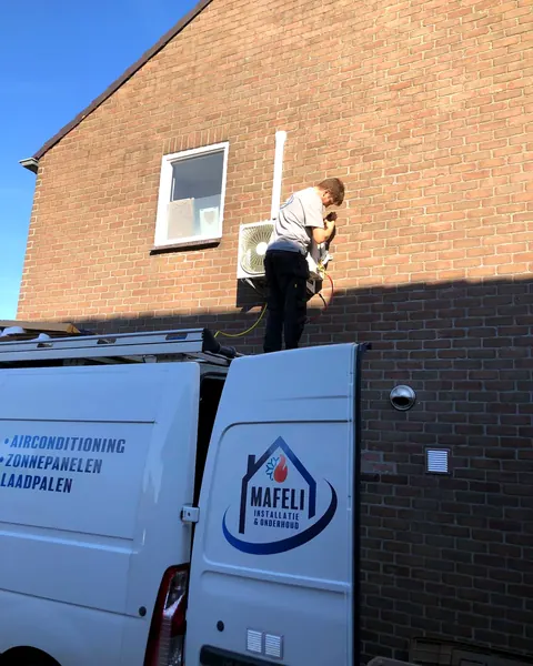 Monteur van Mafeli hangt een airco-buitenunit aan de gevel, met de bedrijfsbus ervoor